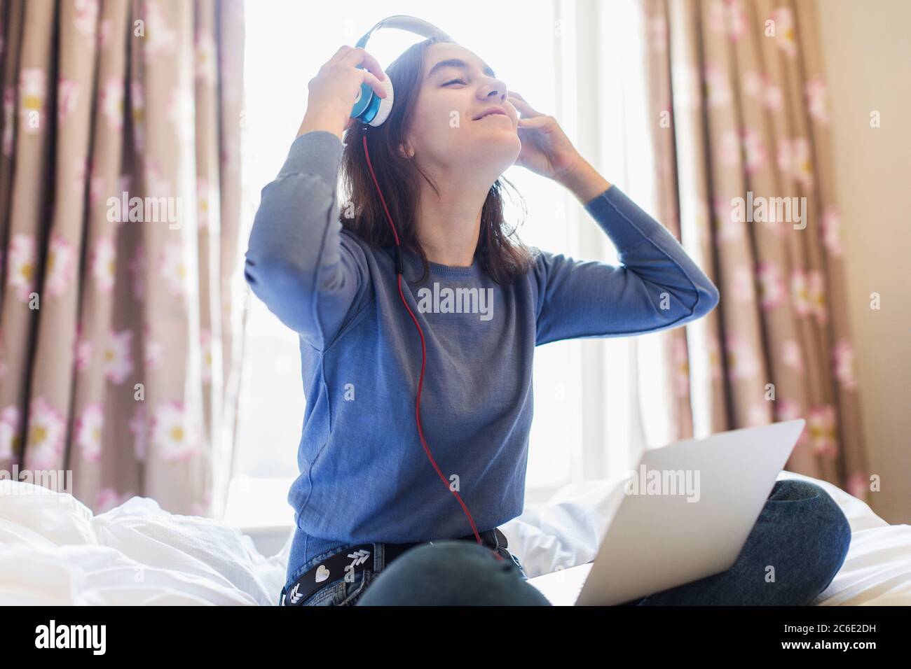 Lächelndes Teenager-Mädchen mit Kopfhörern und Laptop auf sonnigem Bett Stockfoto