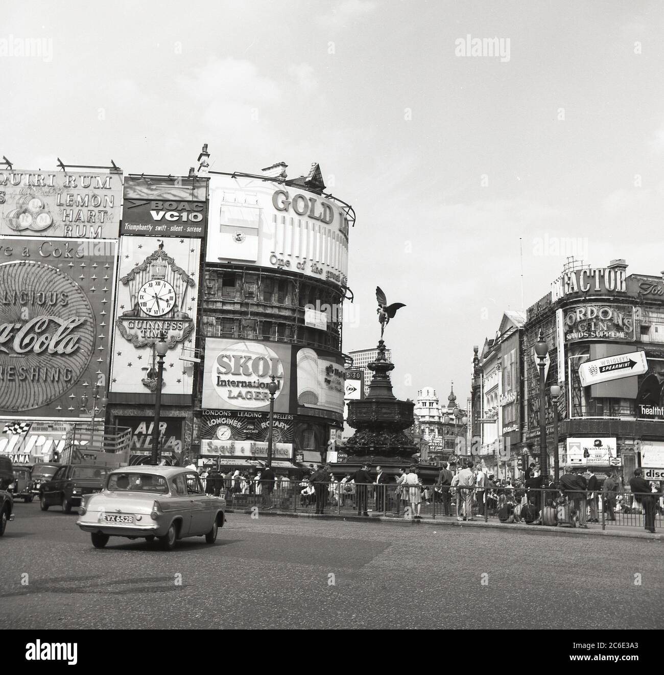 1960er Jahre, historisch, ein Ford Anglia Auto auf dem Piccadilly Circus im Londoner West End, als es ein funktionierender Kreisverkehr war. Die Menschen umgeben den Brunnen der shaftsbury-Gedenkstätte, der gemeinhin als Eros-Statue bekannt ist. Werbetafeln für die großen Marken dieser Zeit sind zu sehen, darunter der BOAC VC10, Skol Lager, Gordons Gin und Wrigleys Kaugummi. Stockfoto