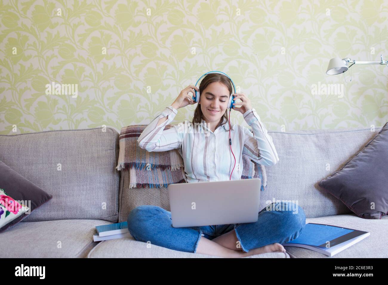 Lächelndes Mädchen im Teenageralter mit Kopfhörern, das auf dem Sofa im Wohnzimmer einen Laptop benutzt Stockfoto