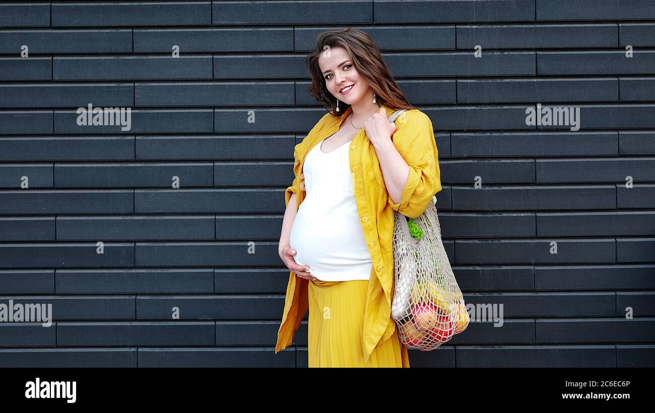 Schwangere Frau in gelber Kleidung mit einer Öko-Tasche mit Obst und Gemüse auf schwarzem Wandhintergrund. Kunststofffreies Konzept Stockfoto