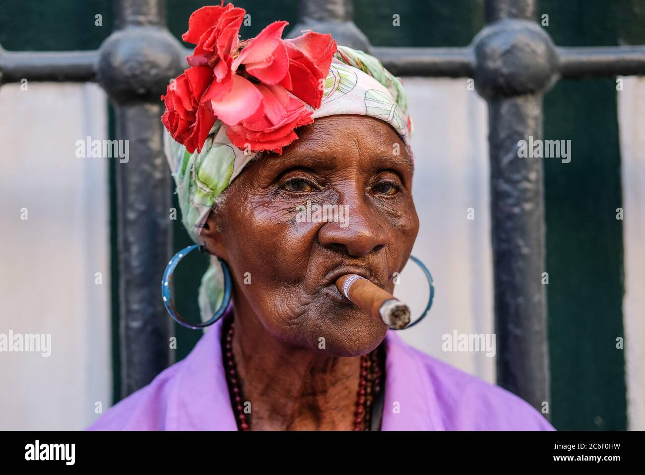 Eine bunt gekleidete Kubanerin raucht eine Zigarre, während sie in Habana Vieja in Havanna, Kuba, an der Straßenseite sitzt. Stockfoto