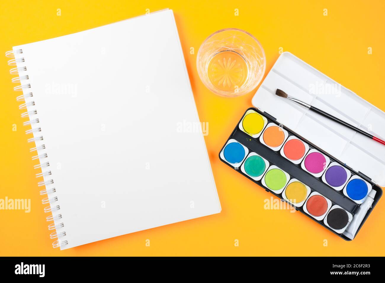 Oben Ansicht von Aquarellpaintbox, Pinsel, Skizzenbuch und Glas Wasser auf orangem Hintergrund, Malerei mit Aquarellen und kreativ sein Stockfoto