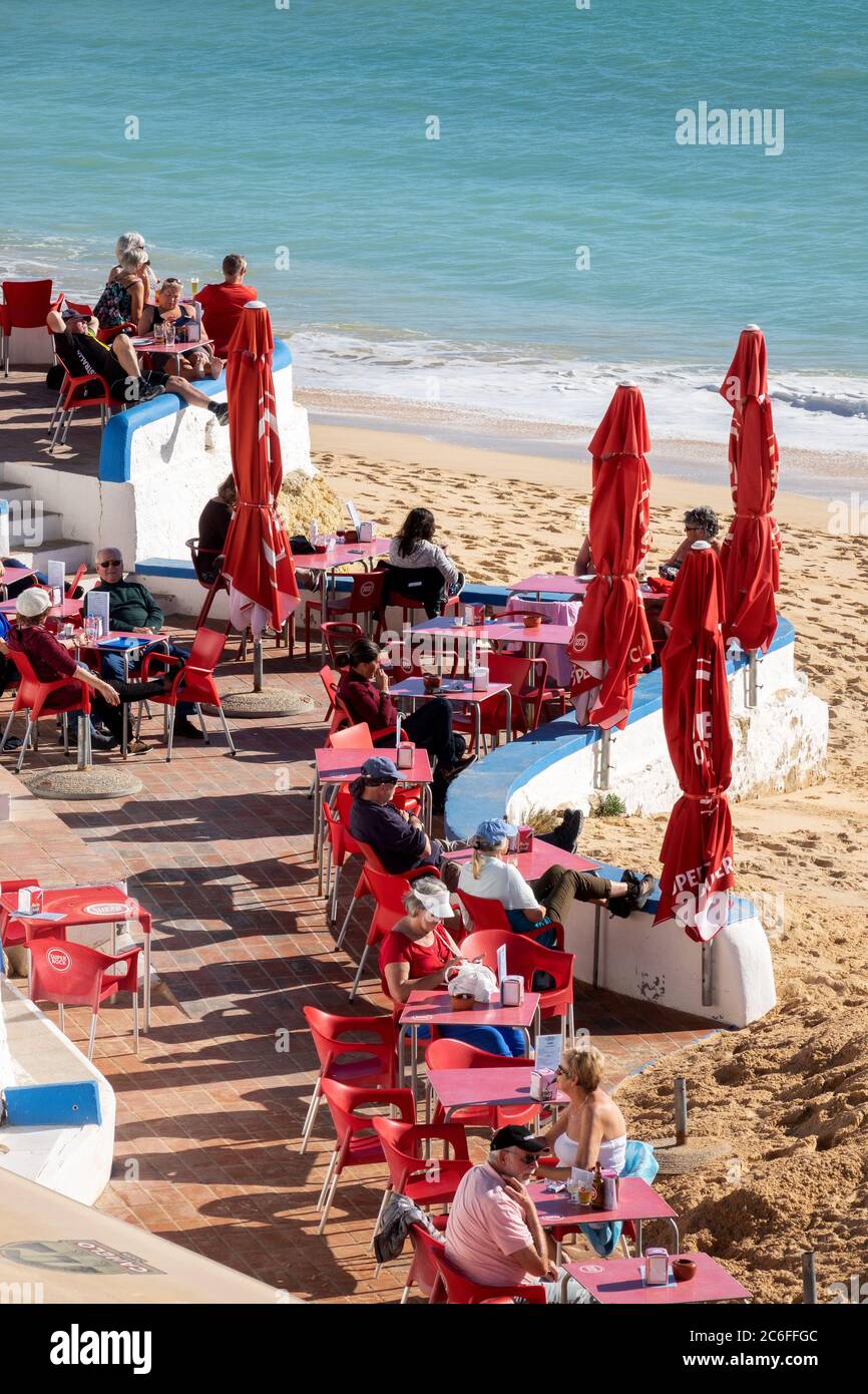 Touristen sitzen in der Wintersonne in der Snack Bar Carlota am Strand in Armacao de Pera an der Algarve Portugal Stockfoto