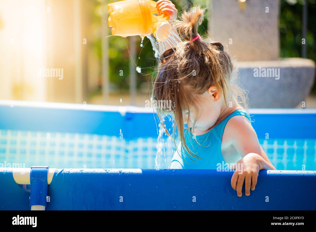 Kleine kaukasische Mädchen, in einem blauen Schwimmbad, erfrischend sich durch Bewässerung ihren Kopf mit einem Spielzeug Dose an einem hellen Sommertag. Kopieren Sie Speicherplatz und su Stockfoto