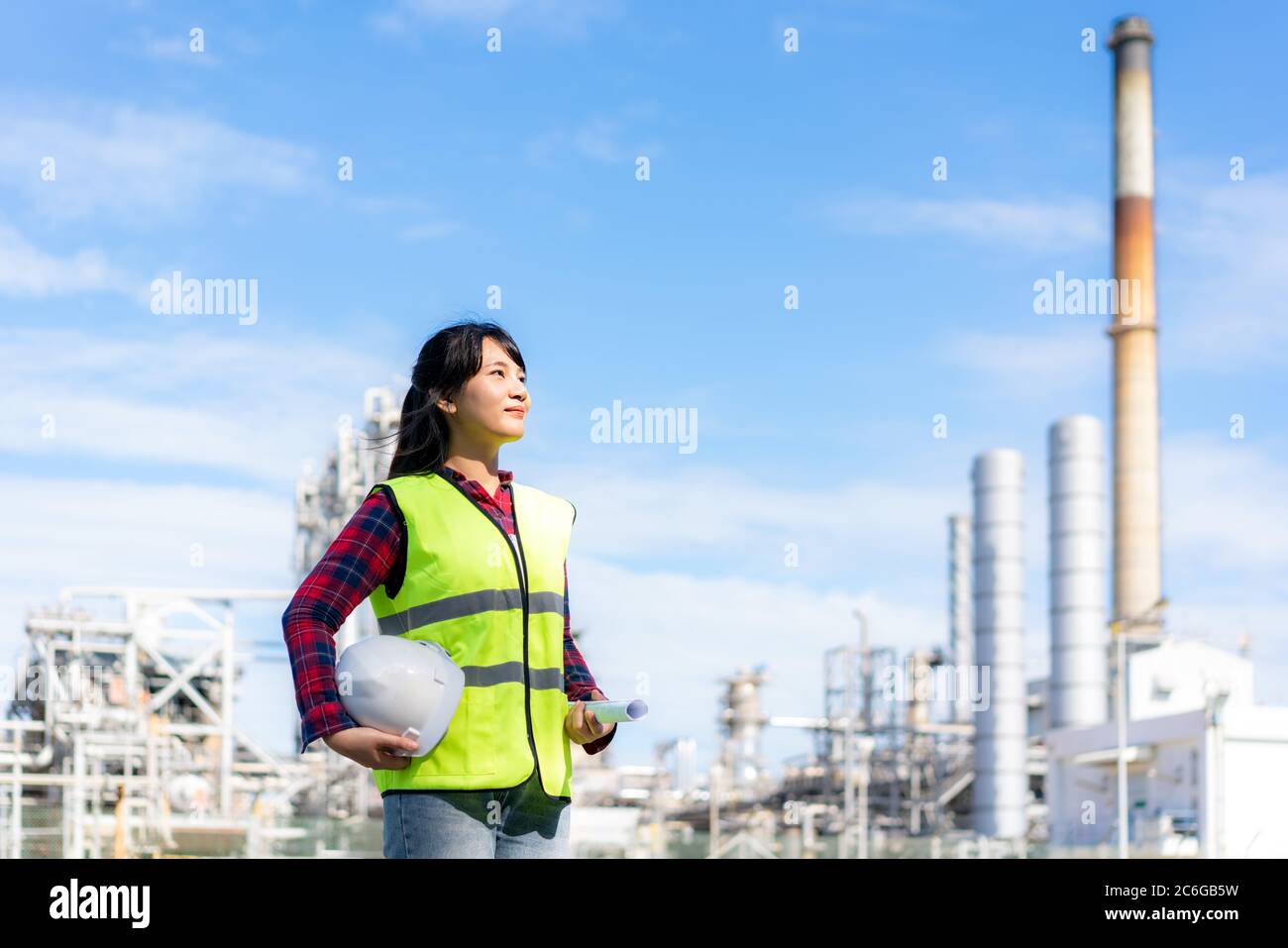 Asiatische Frau Ingenieur mit einem weißen Schutzhelm und Blaupause freuen sich auf die Zukunft mit Ölraffinerie Fabrik im Hintergrund. Stockfoto