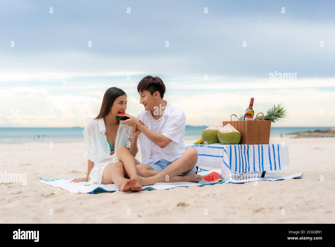 Asiatisches junges Paar, das auf der Picknickdecke sitzt und Wassermelone am Strand und in der Nähe des Meeres mit tropischen Früchten im Hintergrund isst. Sommer, Urlaub, Stockfoto