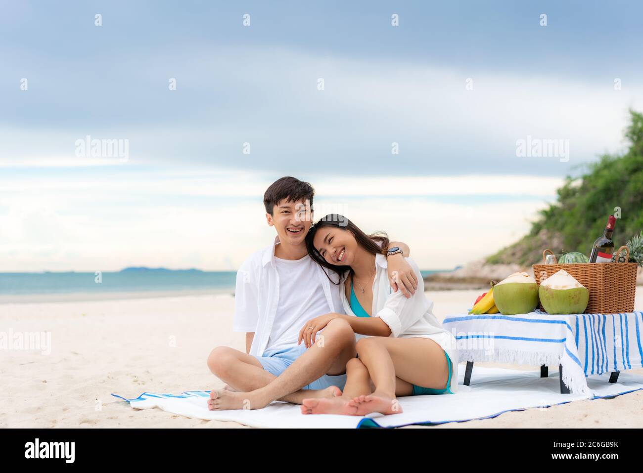 Asiatisches junges Paar sitzt auf der Picknickdecke und entspannen Sie sich am weißen Sandstrand und in der Nähe des Meeres mit tropischen Früchten im Hintergrund. Sommer, Urlaub, V Stockfoto