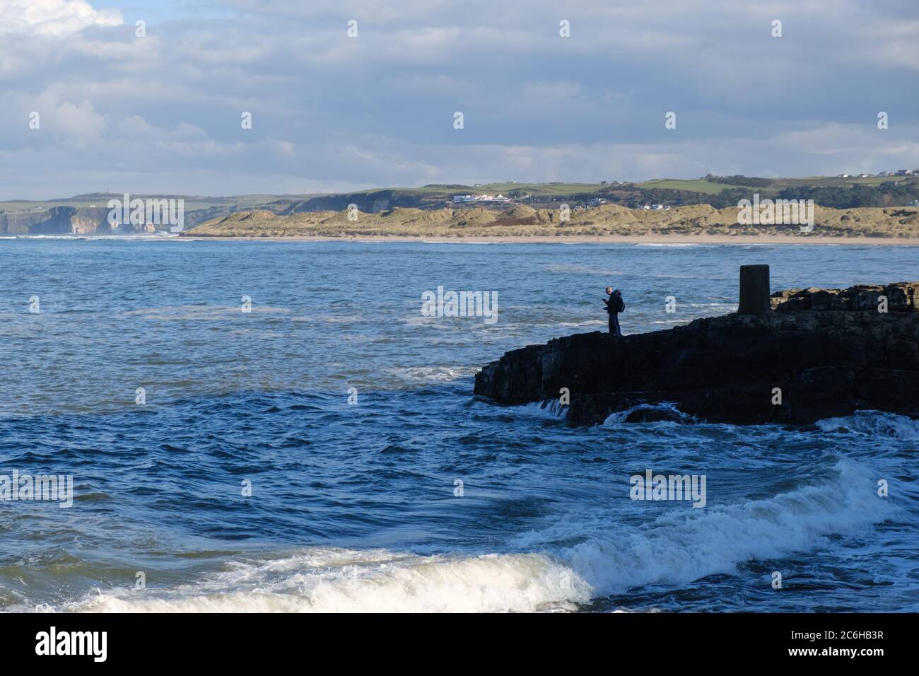 Der junge Mann allein steht an der Küste von Portrush, Co. Antrim, Nordirland verloren in Kontemplation, als er auf sein Telefon herunterschaut Stockfoto