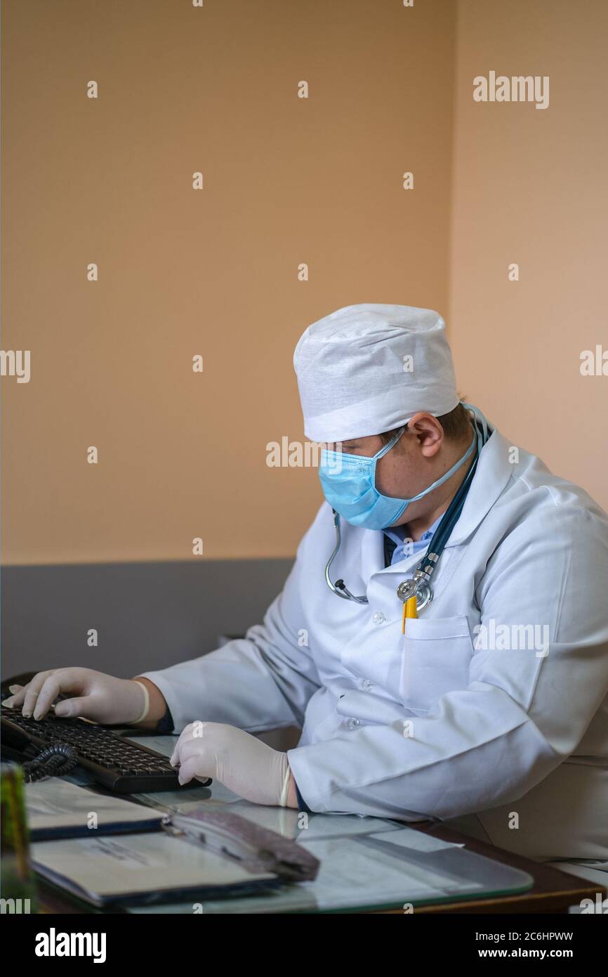 Maskierter Arzt mit Computer. Allgemeinpraktiker arbeitet mit Computer-Anwendung sitzen am Schreibtisch in seinem Büro. Stadtkrankenhaus. Mai 2020, Brovary Stockfoto
