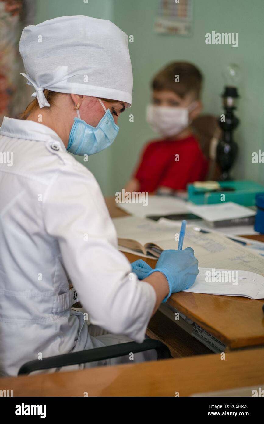 Der Arzt schreibt in der Papierkarte des kleinen Patienten. Maskierter Sanitäter arbeitet in seinem medizinischen Büro. Seitenansicht. Stadtkrankenhaus. Mai 2020, Brovary, Ukraine Stockfoto