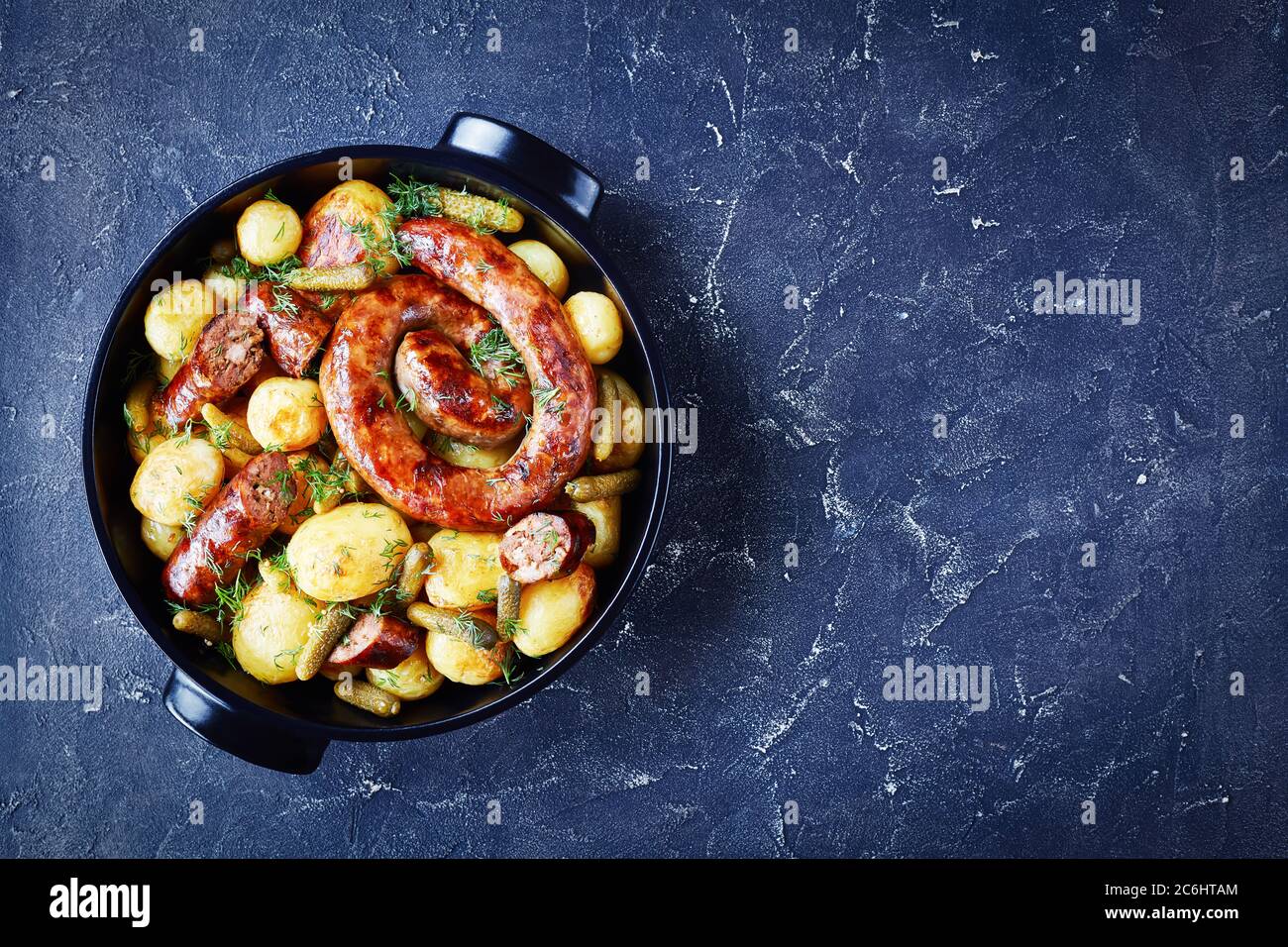 Gebratene neue Kartoffel mit italienischer Schweinewurst auf einem schwarzen Gericht mit Gurken gebraten und mit Dill auf dunklem Betonboden bestreut, Draufsicht, fl Stockfoto
