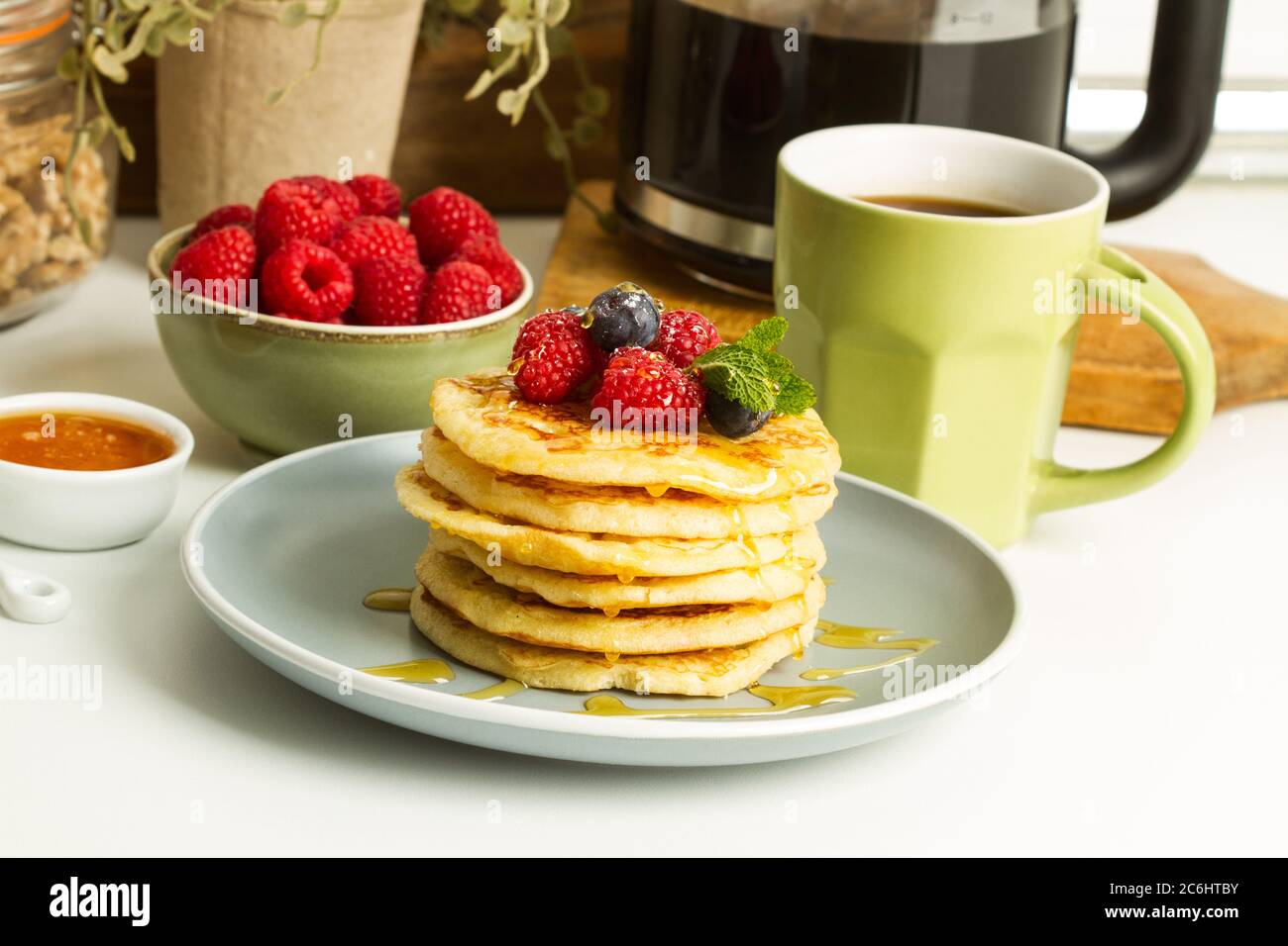 Pfannkuchen mit Beeren und Honig auf einem grauen Teller und mit einer Tasse Kaffee auf einer Küchentheke Stockfoto
