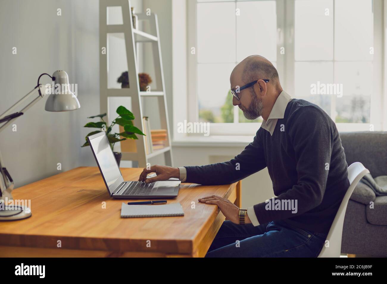 Online arbeiten. Älterer Mann, der Laptop-Text schreibt, während er zu Hause an einem Tisch in einem Raum sitzt. Stockfoto