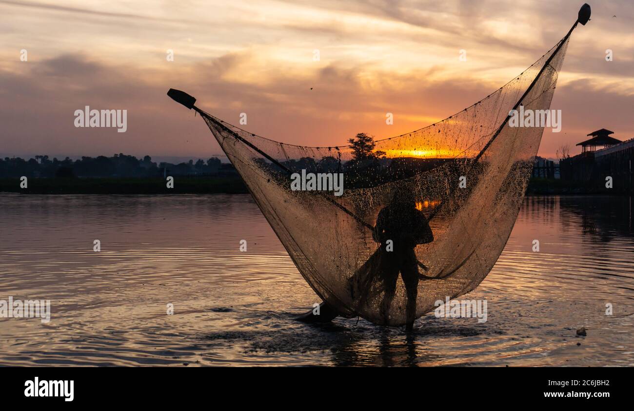 Männer fischen mit dreieckigem Schaufelnetz am frühen Morgen bei Sonnenaufgang in der Nähe der ikonischen U-Bein Brücke, auf flachen See Taungthamanin, Amarapura, Mandalay, Myanmar Stockfoto