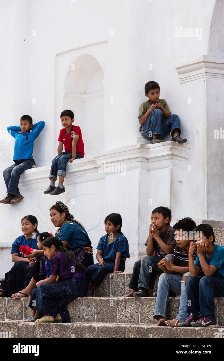 Guatemala, Solola Department, San Antonio Palopo, EINE Cakchiquel Maya-Frau und Kinder beobachten ein Programm von den Stufen der Kirche aus. Die Frau und die Mädchen tragen das traditionelle Kleid von San Antonio Palopo Guatemala, einschließlich der aufwendigen cinta oder Haarwickel, gewebte blaue Huipil Bluse, faja oder Gürtel und corte Rock. Die Jungen tragen alle moderne Kleidung. Stockfoto