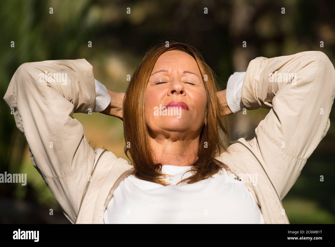 Portrait Attraktive reife Frau sitzen entspannt und glücklich im Freien, mit Armen hinter dem Hals, freundlich, nachdenklich, mit verschwommenem Hintergrund. Stockfoto