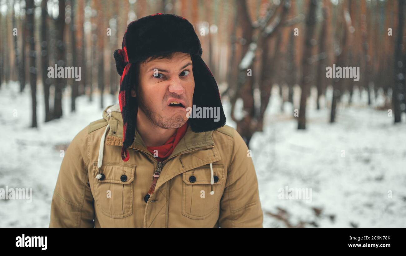 Der Mensch grimaces im Wald in der Wintersaison. Portrait von verrückten Mann tragen Ohrklappe Hut und grimacing Gesicht aus nächster Nähe im Winterwald. Stockfoto