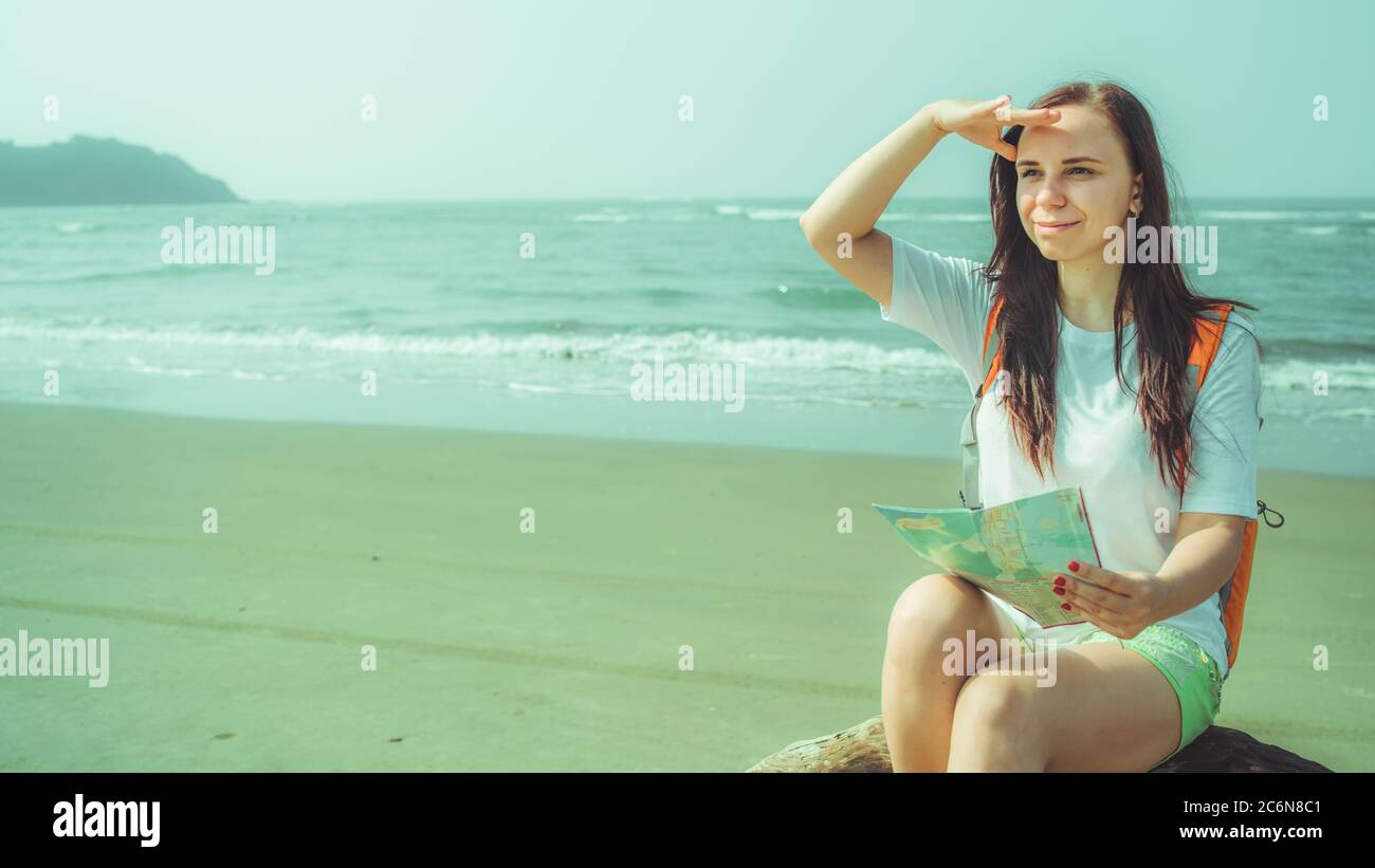Weibliche Touristen lesen Karte in der Nähe des Meeres. Barfuß Frau mit Rucksack sitzt auf Treibholz und untersuchen Karte während auf Sandstrand in der Nähe winkende Meer ruhen Stockfoto