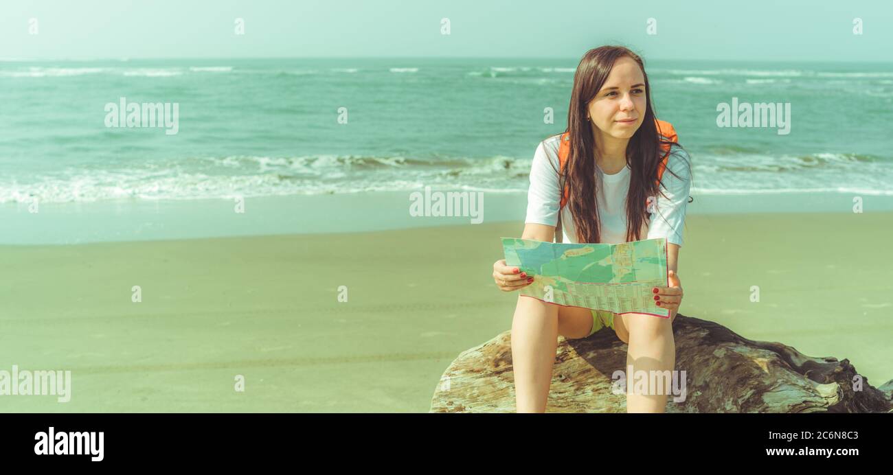 Weibliche Touristen lesen Karte in der Nähe des Meeres. Barfuß Frau mit Rucksack sitzt auf Treibholz und untersuchen Karte während auf Sandstrand in der Nähe winkende Meer ruhen Stockfoto