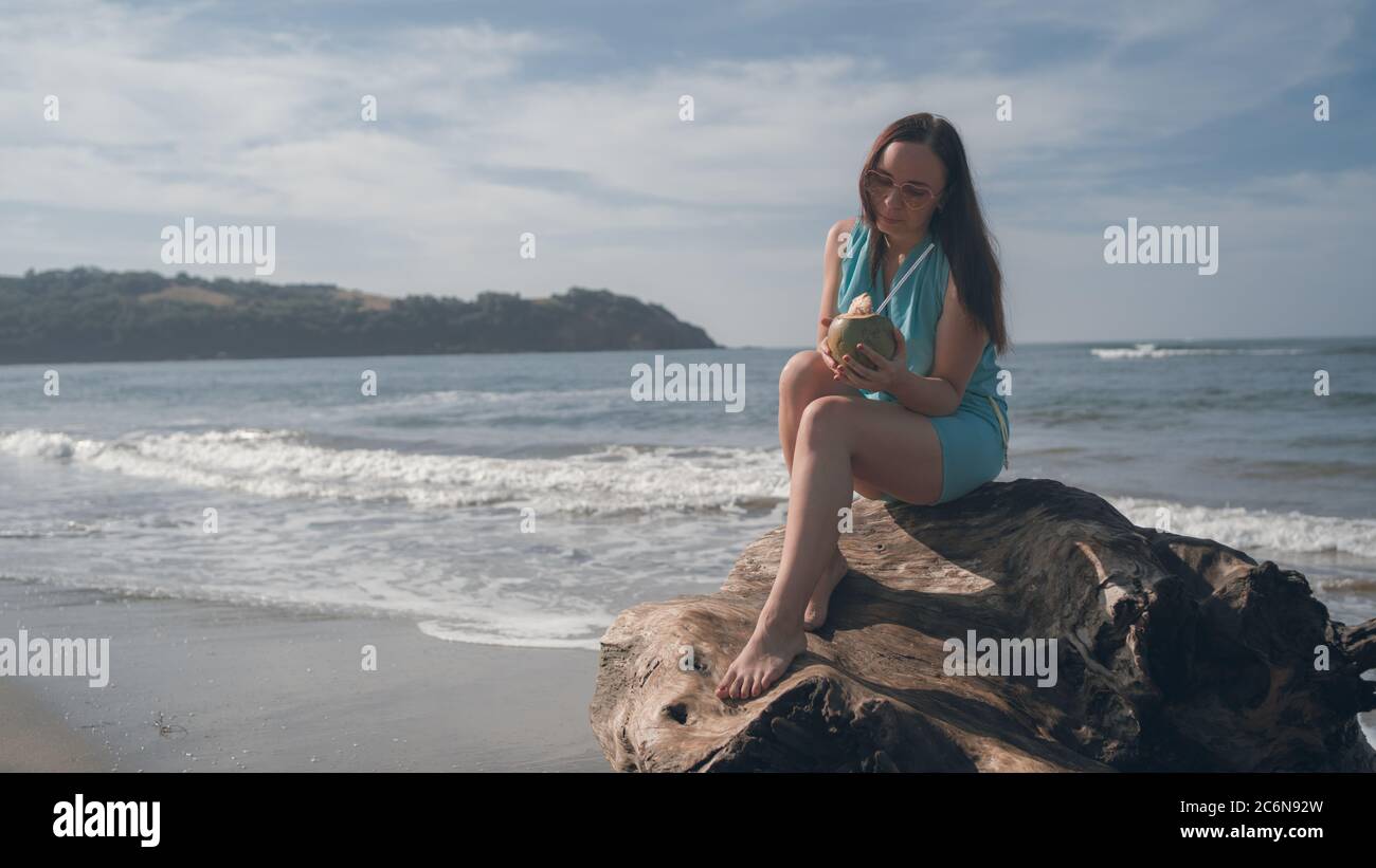 Eine schöne junge Frau sitzt auf einem Baumstamm und trinkt die Kokosnuss am Meer oder Meer an einem hellen sonnigen Tag. Stockfoto