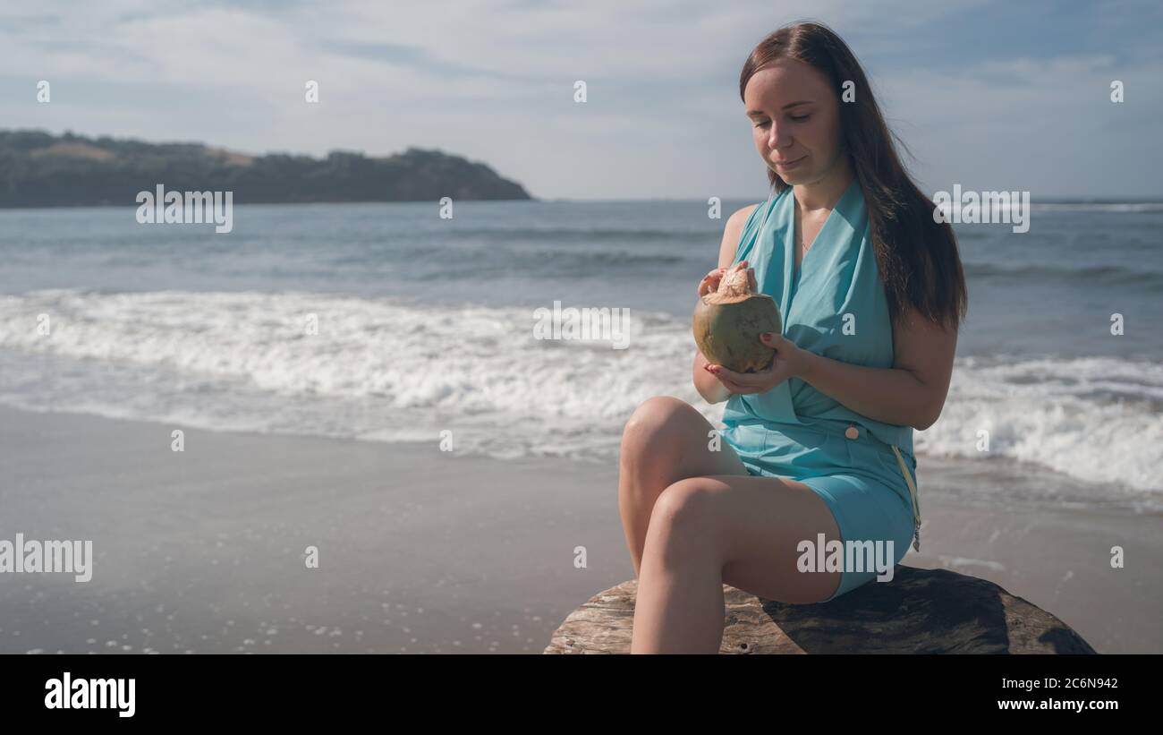 Eine schöne junge Frau sitzt auf einem Baumstamm und trinkt die Kokosnuss am Meer oder Meer an einem hellen sonnigen Tag. Stockfoto