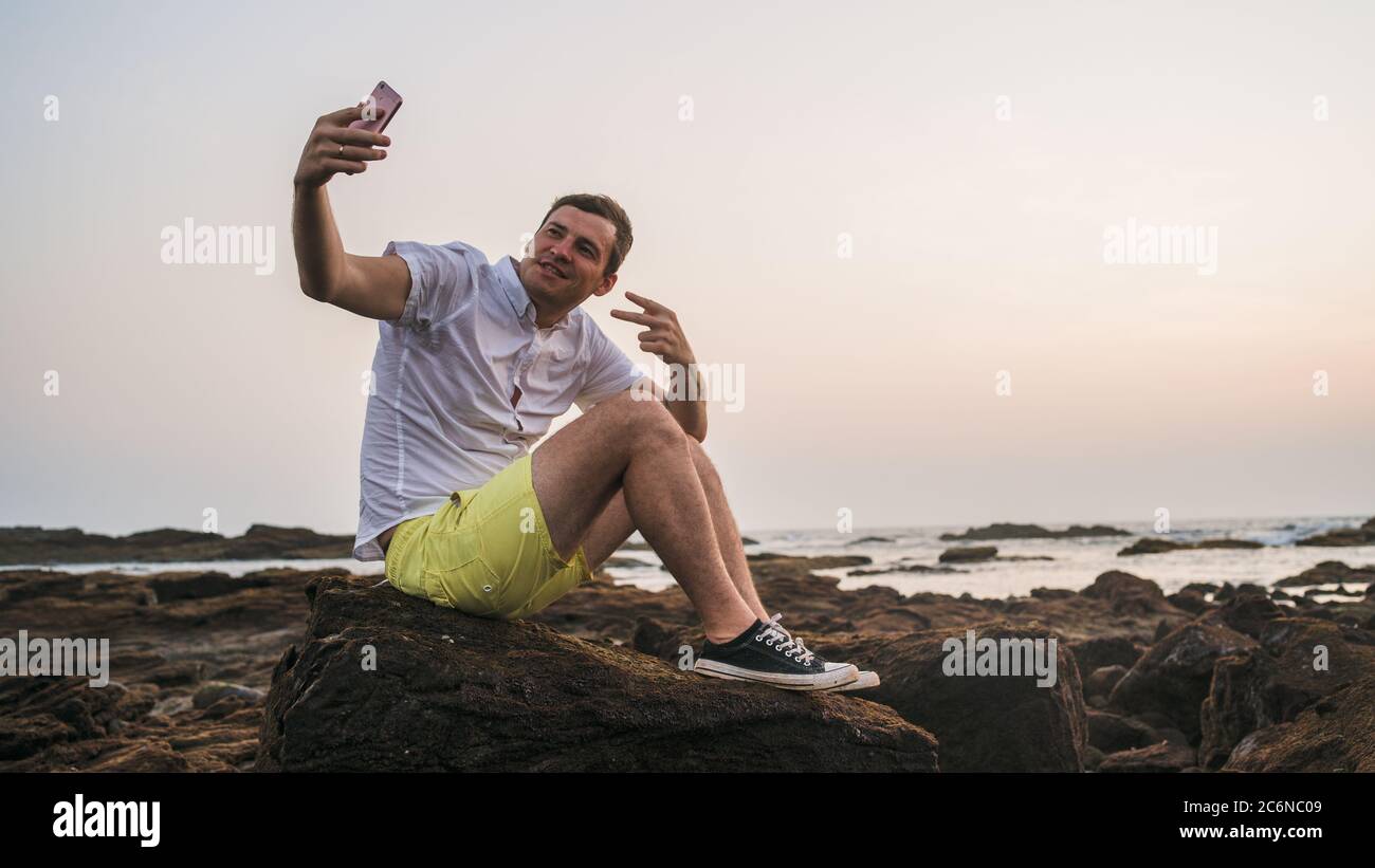 Entspannter Mann sitzt auf Stein und Selfie auf dem Smartphone am Ufer. Seitenansicht des angenehmen Mannes, der Zeit verbringt, den Urlaub zu genießen und zu fotografieren Stockfoto