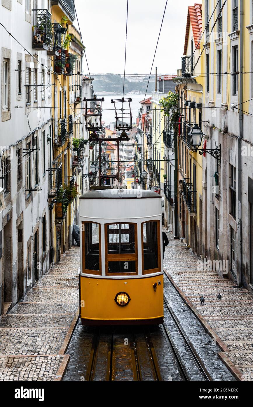 Traditionelle gelbe Straßenbahn in einer engen Straße in Lissabon an einem regnerischen Wintertag, mit dem Fluss Tejo im Hintergrund entkokend. Stockfoto