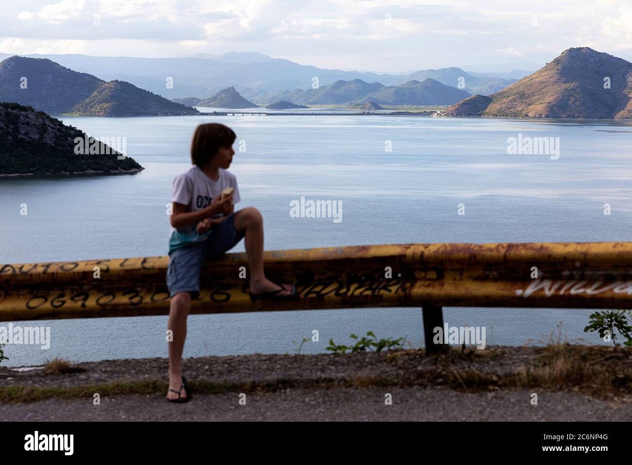 Junge sitzt am Straßenzaun mit einem Snack mit einem schönen Blick über den See Skadar und Virpazar Brücke bei Sonnenuntergang, Montenegro Stockfoto