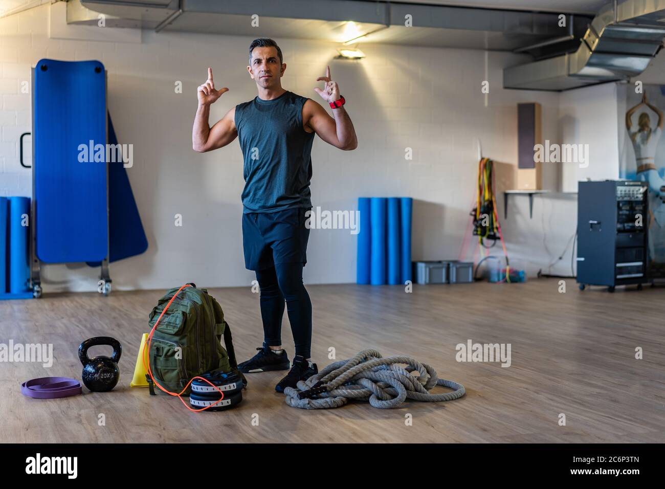 Motivierter Boot Camp Trainer steht mit Fitnessgeräten in der Turnhalle. Hanteln, Seil, Sandsack auf Holzboden. Portrait des Trainers mit den Muskeln Stockfoto