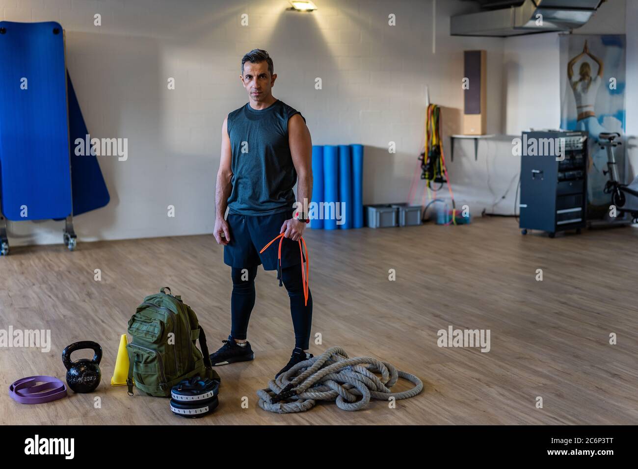 Motivierter Boot Camp Trainer steht mit Fitnessgeräten in der Turnhalle. Hanteln, Seil, Sandsack auf Holzboden. Porträt des libanesischen Sportlehrers. Stockfoto