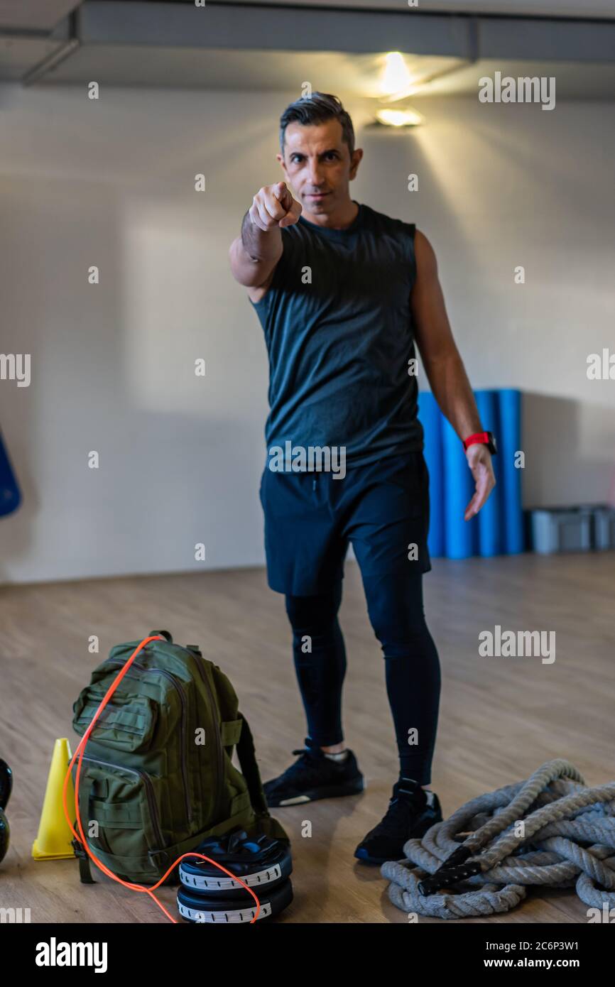 Der Trainer zeigt mit dem Zeigefinger auf die Kamera. Motivierter Boot Camp Instructor steht mit Hanteln, Seil, Sandsack auf Holzboden in der Turnhalle. Stockfoto