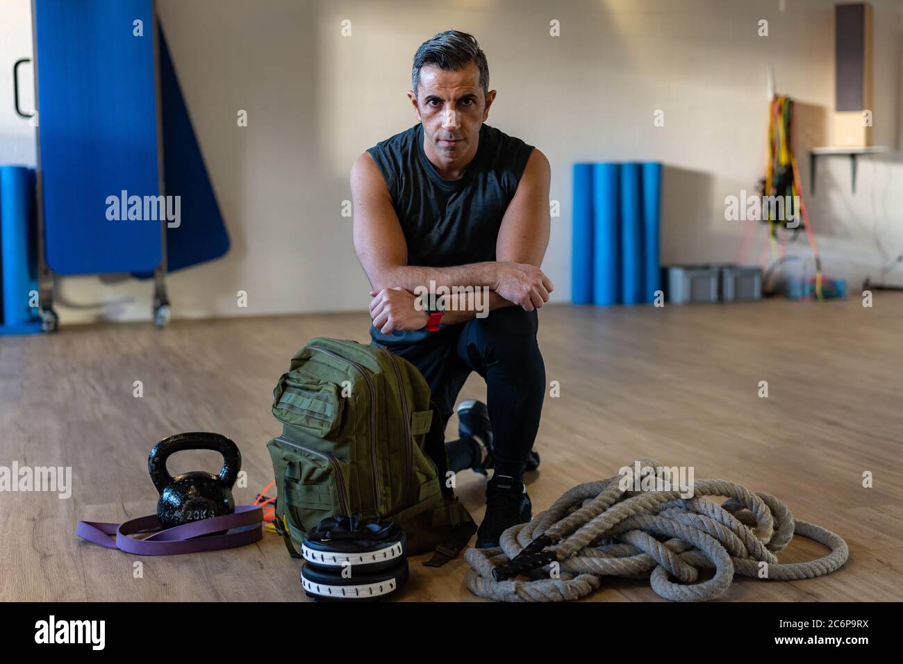 Motivierter Boot Camp Trainer kniet mit Fitnessgeräten in der Turnhalle. Hanteln, Seil, Sandsack auf Holzboden. Porträt des libanesischen Sportlehrers. Stockfoto