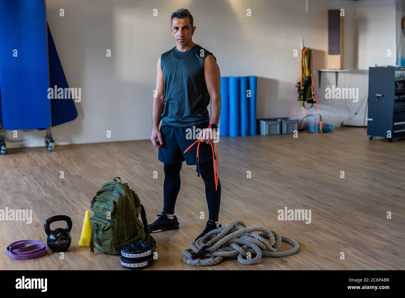 Motivierter Boot Camp Trainer steht mit Fitnessgeräten in der Turnhalle. Hanteln, Seil, Sandsack auf Holzboden. Porträt des libanesischen Sportlehrers. Stockfoto