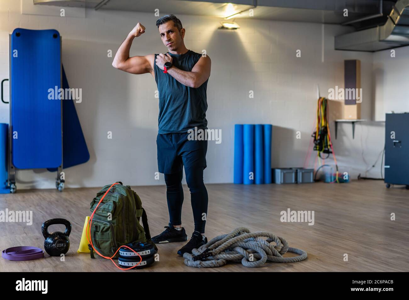 Fitnesslehrer zeigt Muskeln. Motivierter Boot Camp Trainer steht mit Fitnessgeräten in der Turnhalle. Hanteln, Seil, Sandsack auf Holzboden. Innen. Stockfoto