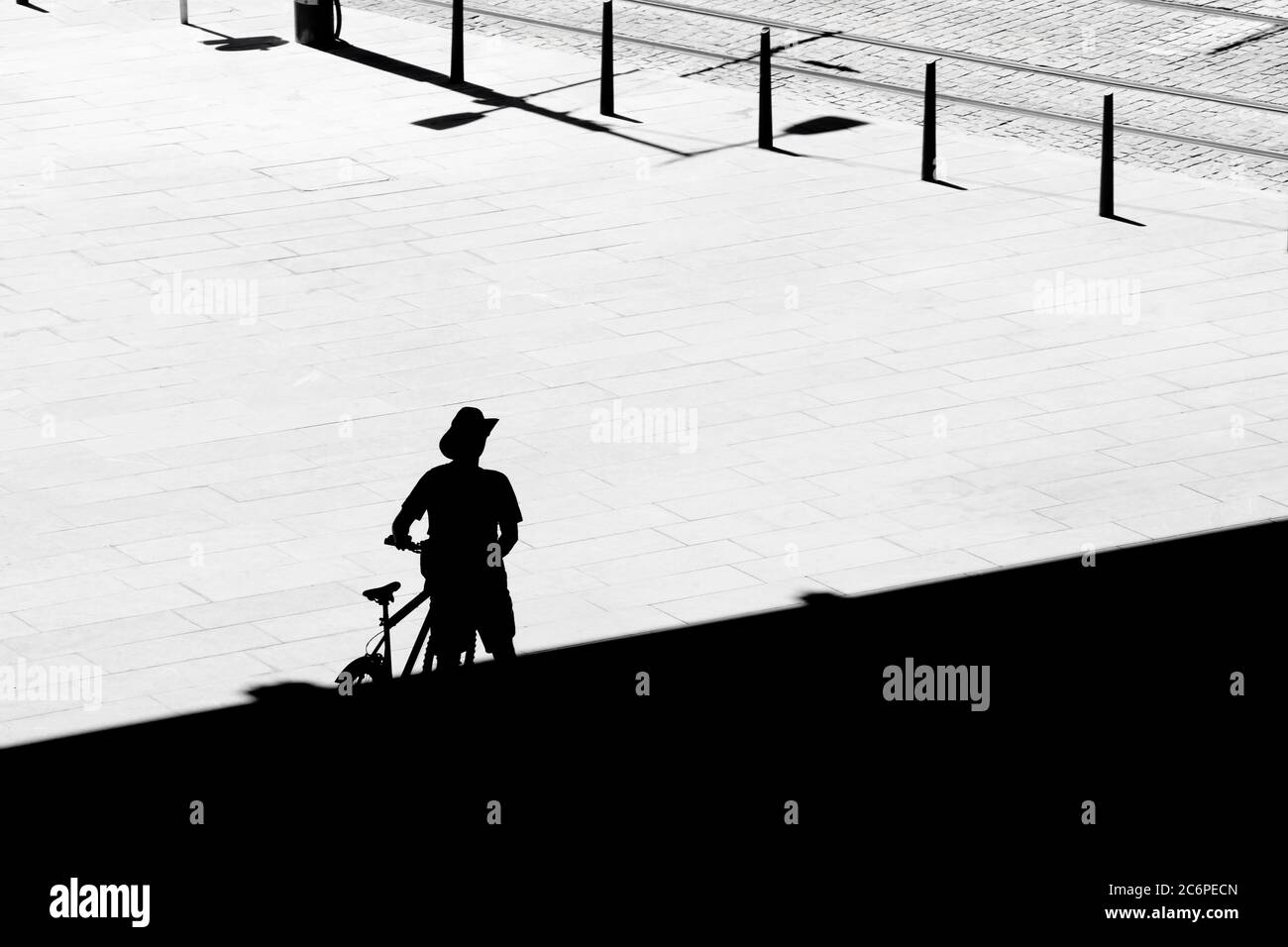 Silhouette eines einsamen Mann zu Fuß aus dem Schatten neben seinem Fahrrad während der Überquerung des Stadtplatzes an einem heißen Sommertag , hochwinkel Rückansicht Stockfoto