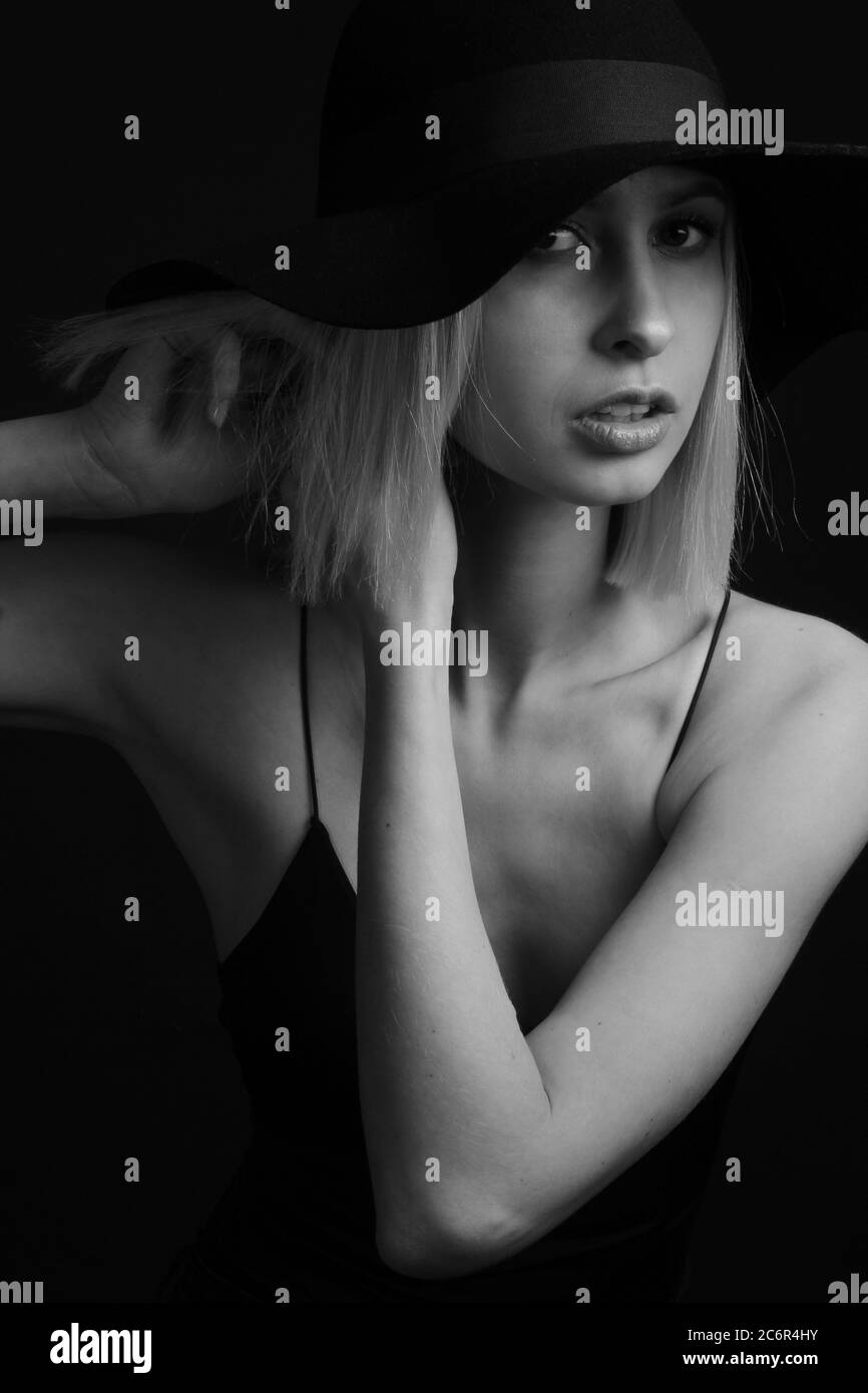 Schwarz-weiße Portrait Blondinen auf schwarzem Hintergrund in einem Hut und Tank Top mit dünnen Trägern. Hände am Ohr, passt den Ohrring Stockfoto