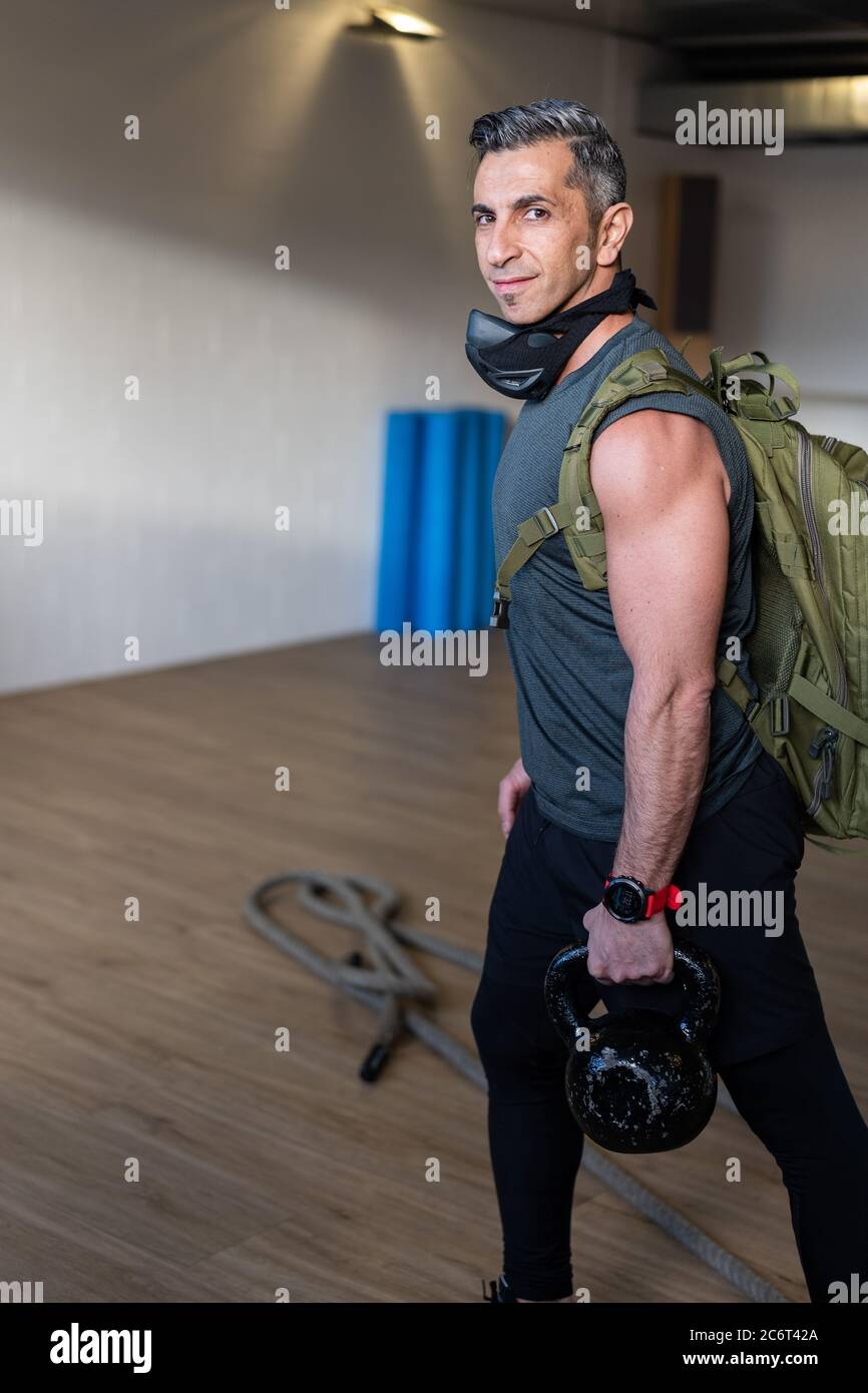 Motivierter Boot Camp Trainer steht mit Fitnessgeräten in der Turnhalle. Tragen Sie Trainingsmaske, Hanteln und Sandsack, Seil auf Holzboden. smi-Instruktor Stockfoto