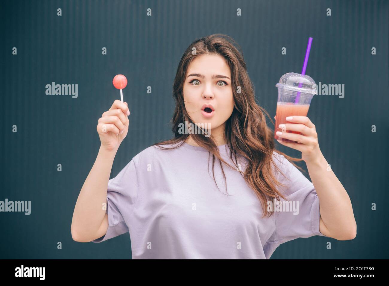Junge stilvolle trendige Frau isoliert über graublauen Hintergrund. Emotionale Mädchen hält Limonade in Kunststoff-Tasse und Lollipop in einer anderen Hand. Erstellen von emo Stockfoto
