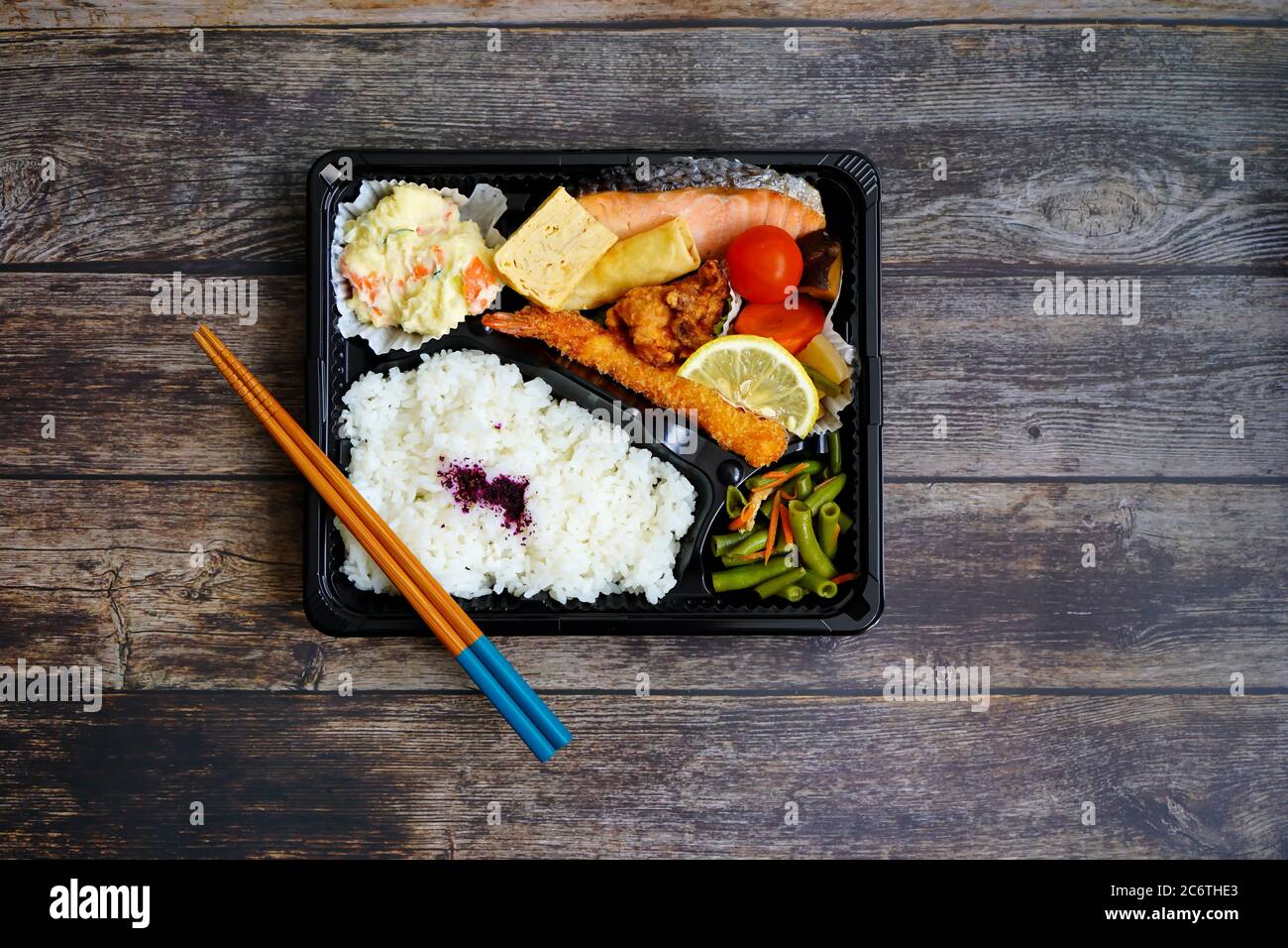 Bunte japanische „Makunouchi“ Bento-Box mit Lachs, gebratenen Garnelen, Kartoffelsalat, Tofu, gegrilltem Hähnchen, Reis und Gemüse. Rustikaler dunkler Holztisch. Stockfoto