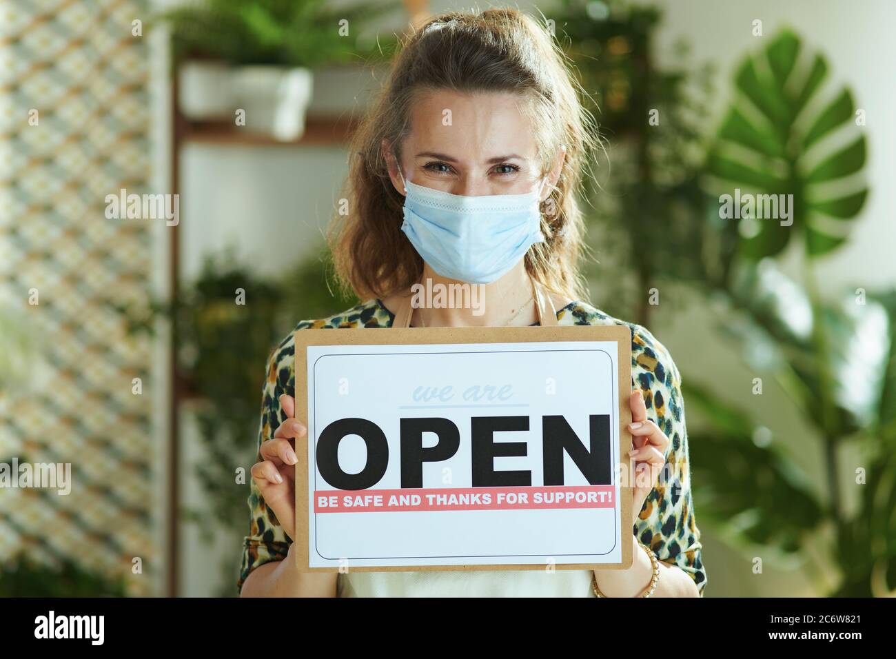 Überleben von Kleinunternehmen nach einer Pandemie des Covid-19. Portrait von lächelnd trendy 40 Jahre alte Kleinunternehmen Besitzerin Frau in Schürze mit medizinischen Maske zeigt Stockfoto