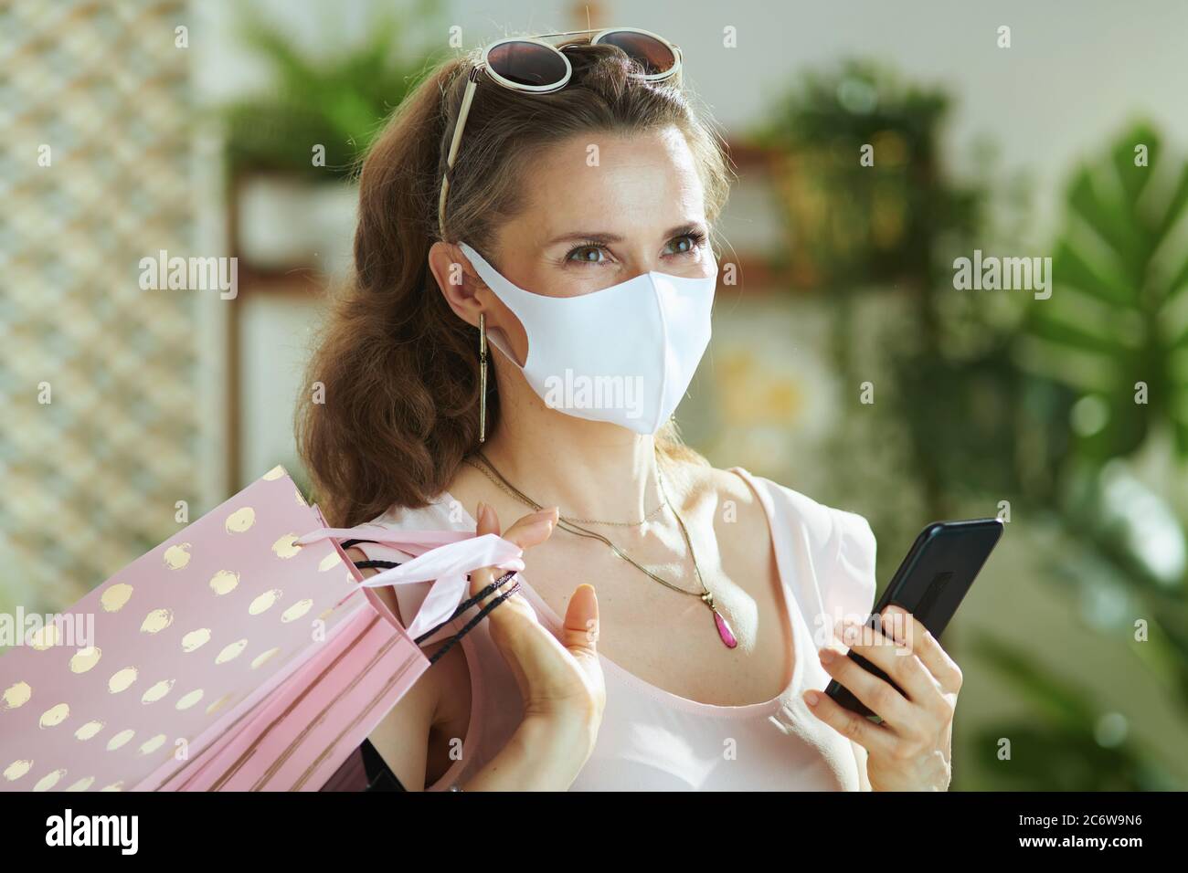 Leben während der Coronavirus-Pandemie. Trendige Frau Shopper in rosa Bluse mit medizinischen Maske, Papier Einkaufstaschen und Smartphone-Kauf im Internet. Stockfoto