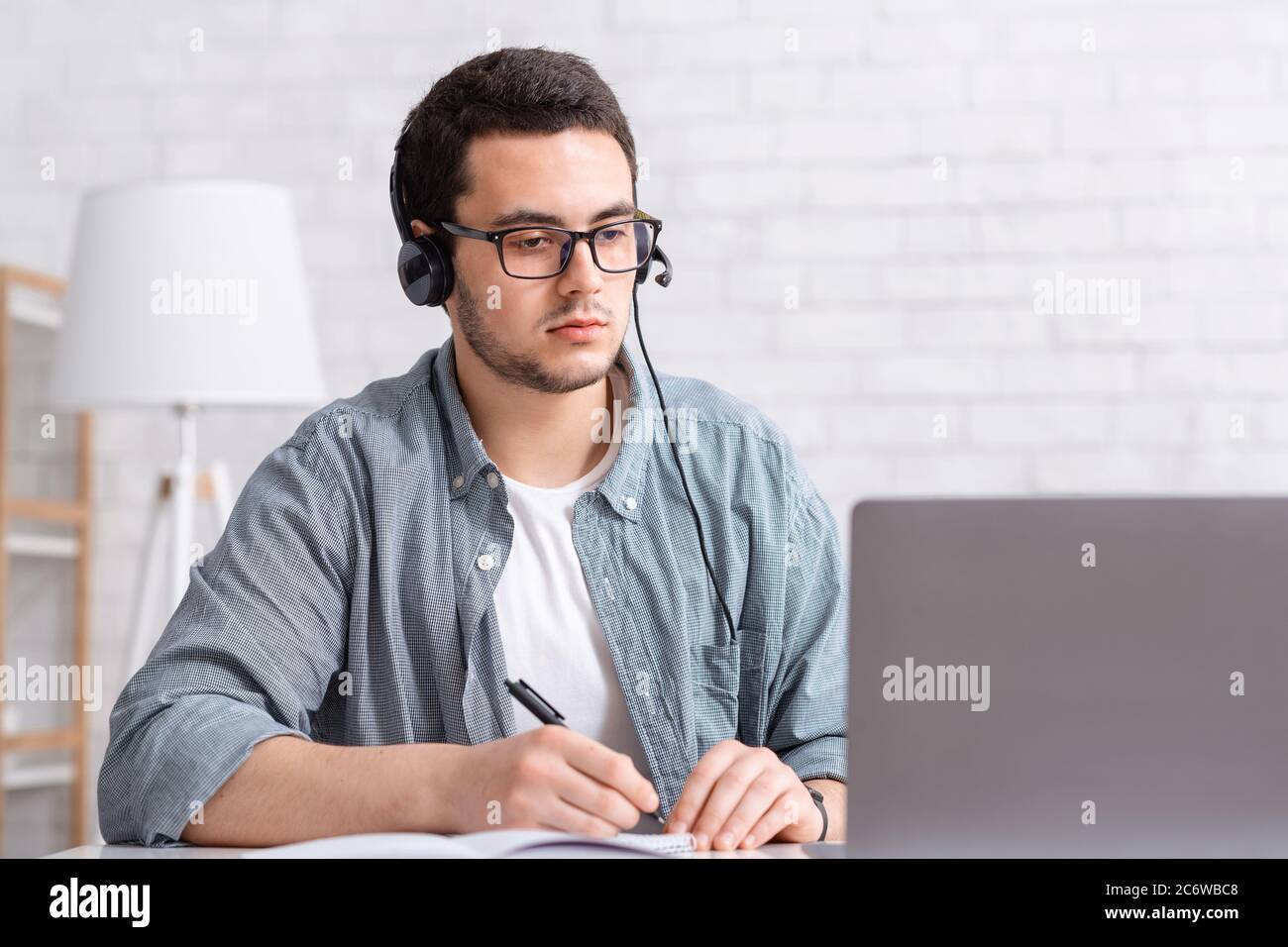 Webinare und Schulungen. Fokussierter Kerl mit Brille und Kopfhörer macht Notizen in Notebook und sieht Laptop Stockfoto