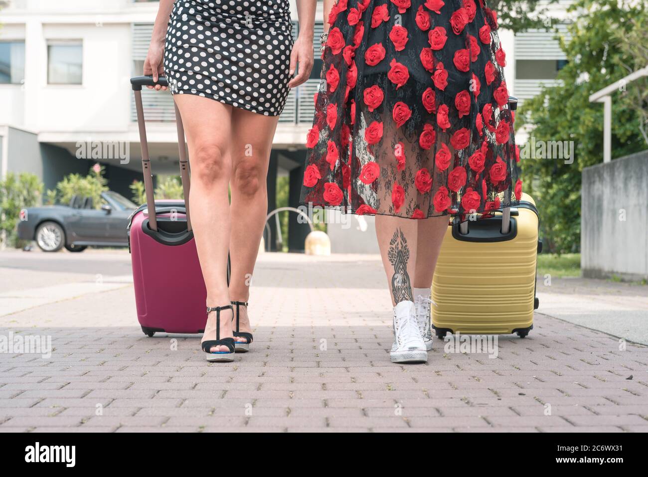 Von unten zugeschnittenes Foto von zwei eleganten Touristenfrauen, die mit Koffern auf der blumigen Straße der Stadt spazieren. Stockfoto