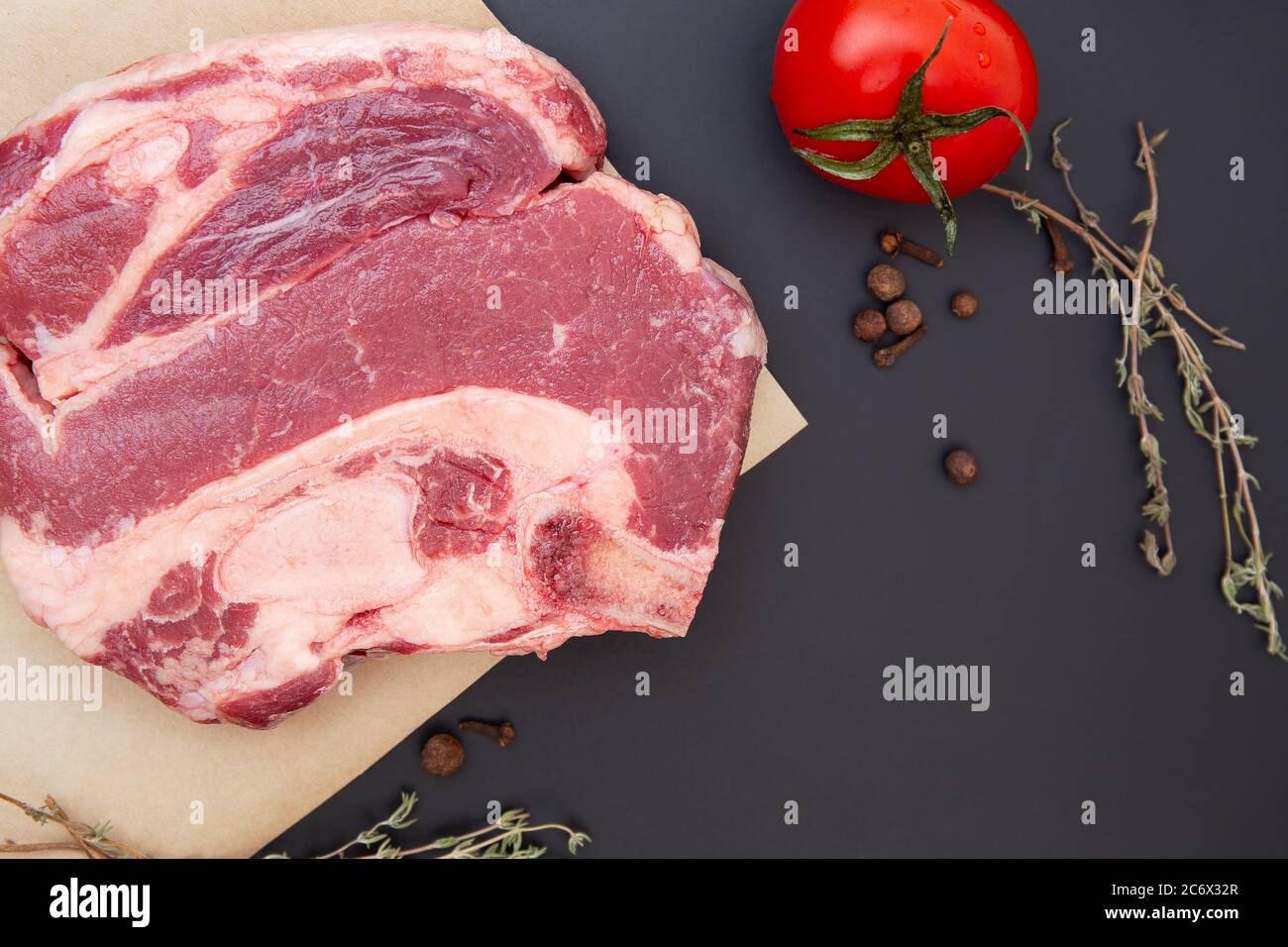 Frisches rohes Rindersteak mit Gewürzen, Kräutern und Gemüse. Das Fleisch ist auf dem Knochen. Thymian, schwarzer Pfeffer, Tomaten, Rindfleisch auf schwarzem Stein. Hintergrund für das Essen. Stockfoto