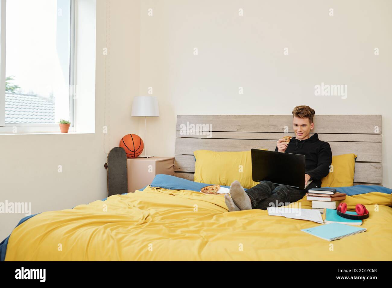 Positive High School Student sitzt auf dem Bett, Pizza essen und Online-Webinar ansehen, wenn zu Hause bleiben während Quarantäne Stockfoto