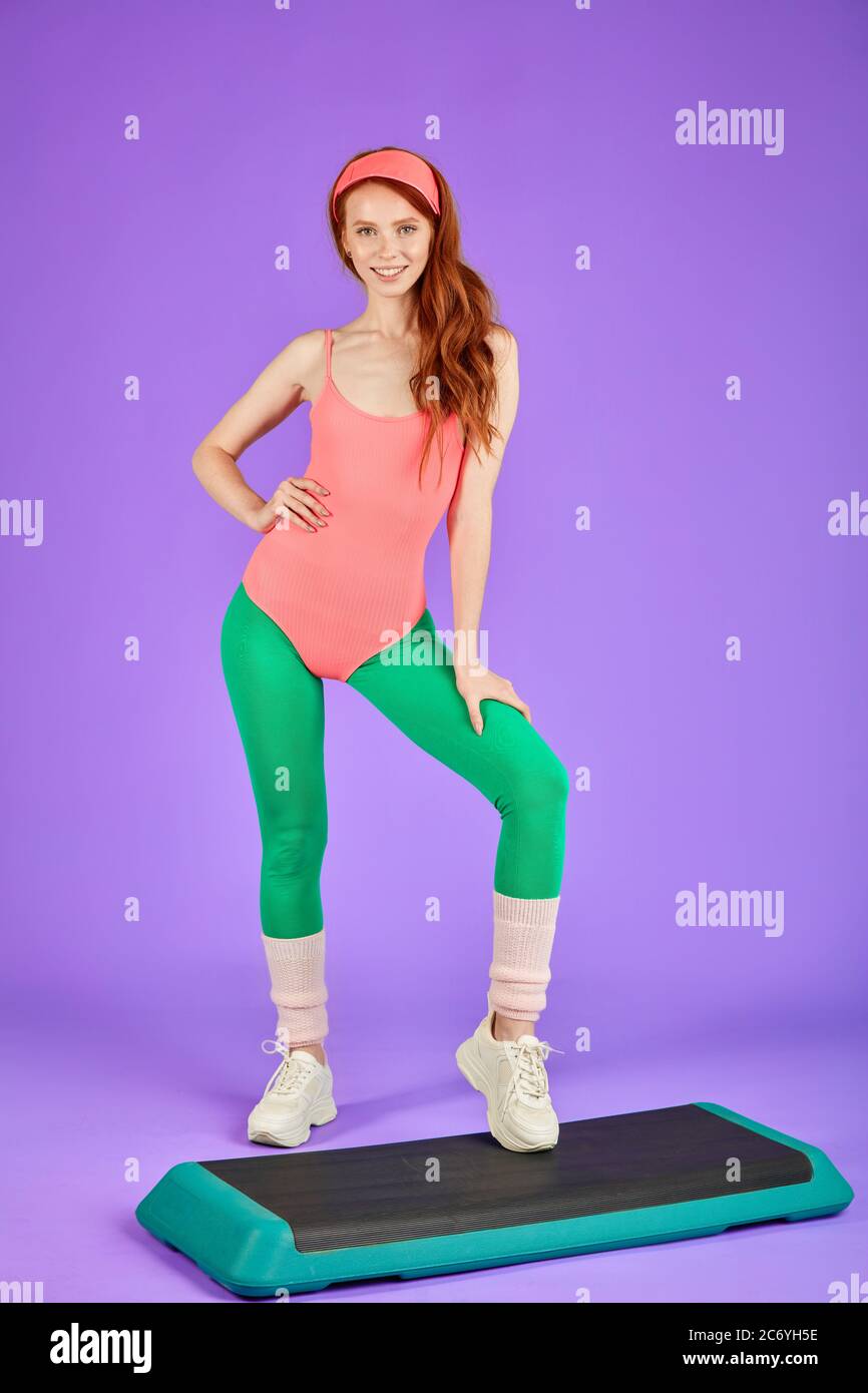 Ginger Mädchen mit perfekter Körperform und lange rote Haare steht auf Schritt während der Pause bei Aerobic-Klasse, trägt 80er Jahre Retro-Stil rosa Bodysuit, Leggins und Stockfoto