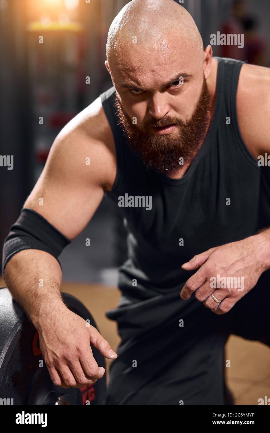 Brutal schöner Athlet, der mit strengen Augen auf die Kamera schaut, in schwarzem Singlet und Shorts gekleidet, auf dem Boden sitzend, Kraft und Stärke ausdrückt Stockfoto