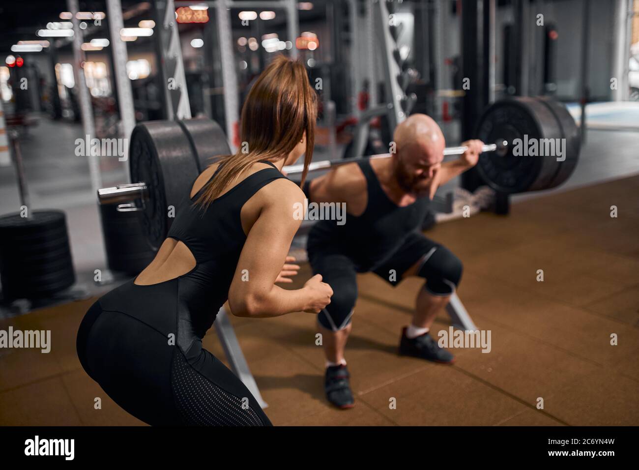 Nahaufnahme einer sportlich-fitter Frau, die mit dem Rücken zur Kamera steht, den brutalen Powerlifter unterstützt und mit schwerer Langhantel auf den Schultern steht, um zu üben Stockfoto