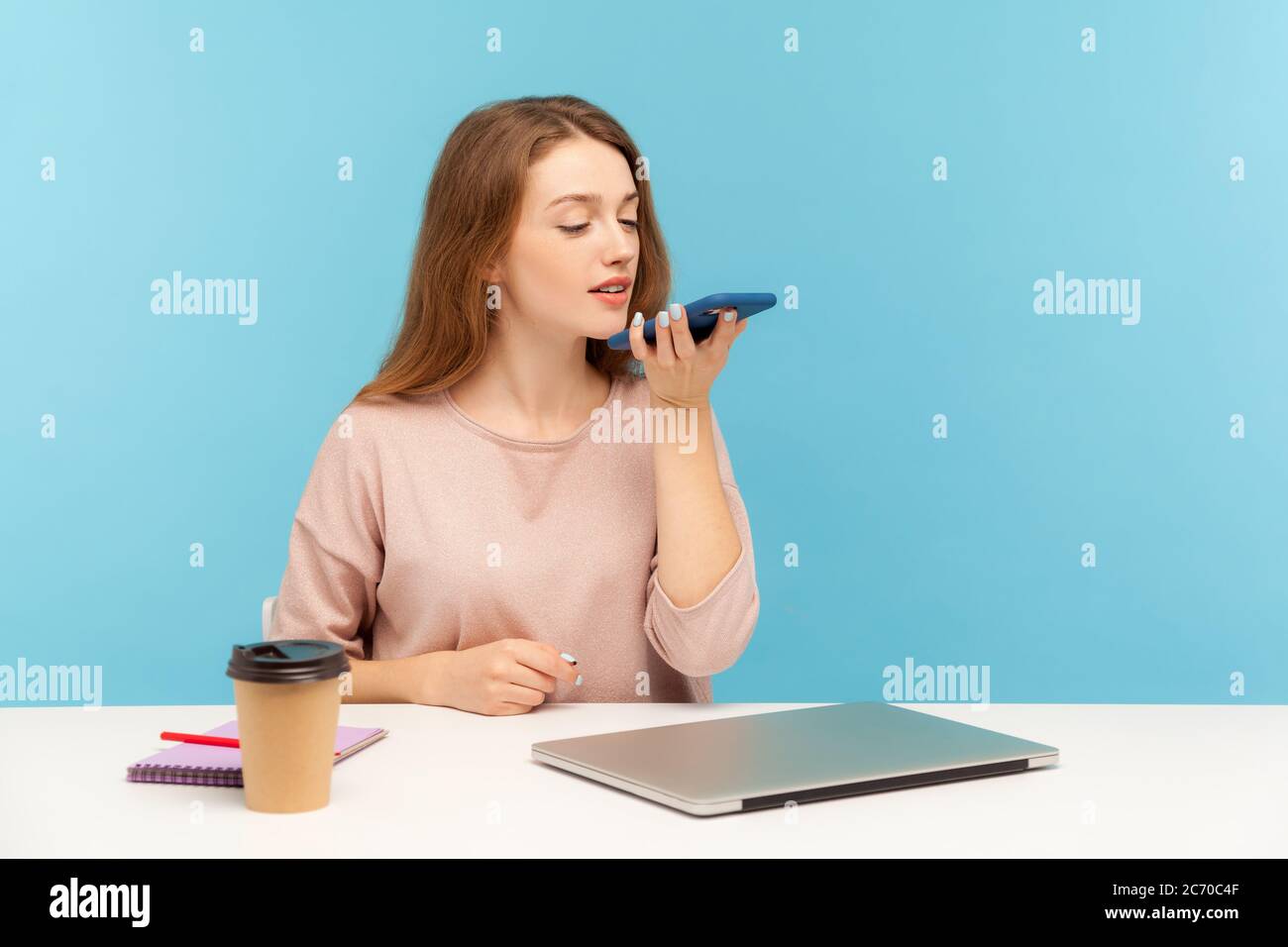 App für digitale Lautsprecher. Junge Angestellte, die am Arbeitsplatz sitzt und mit dem Mobiltelefon spricht, mit dem Sprachassistenten Erinnerungen an ein Geschäftsereignis aufzeichnet Stockfoto