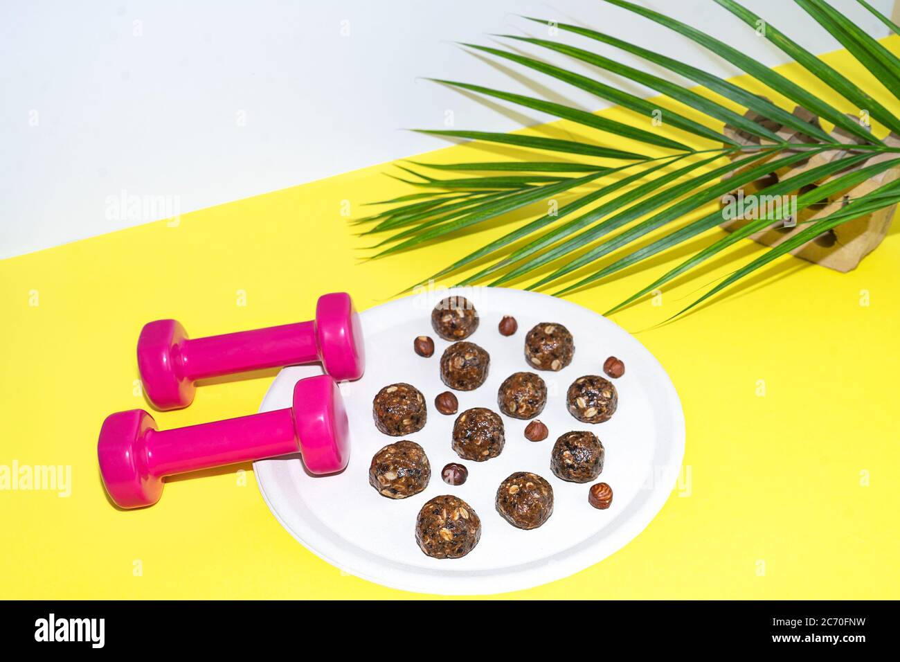 Protein Erdnussbutter Energie Bisse oder Energiebälle. Hausgemachte rohe Schokoladentrüffel mit Nüssen auf Teller mit Hanteln und Palmblatt auf gelbem Hintergrund. Stockfoto